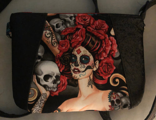 A black shoulder bag with a Day of the Dead sugar skull and red roses print, resting on a light-colored chair with a wooden floor visible in the background.